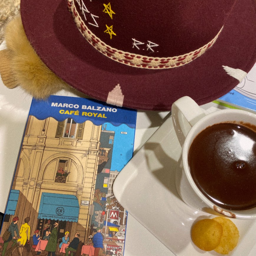 Libro dal titolo Cafè Royal appoggiato accanto a una tazza di cioccolata calda e un cappello